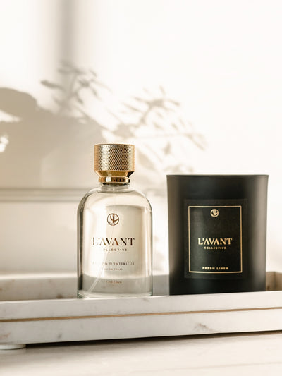 Clear glass bottle with L'AVANT logo stamped in gold foil. Glass bottle with gold topper. Black candle in glass jar with L'AVANT Collective logo. Both products are displayed on a natural color marble tray.