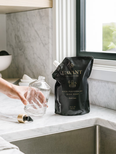 Clear glass custom bottle with brush gold neck on a black pump laying on the bottom left. Hand is holding empty glass bottle tipping towards the black refill dish saop bags with a white cap. All sitting on a marble countertop in kitchen.