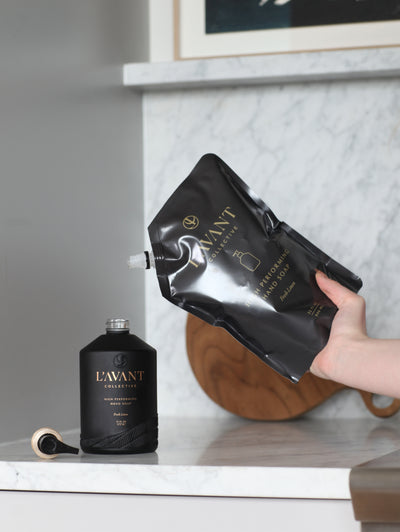 Black custom glass hand soap bottle being filled from a black 32 oz refill bag in hand from the right side.  All resting on a marble counter with marble wall backsplash.