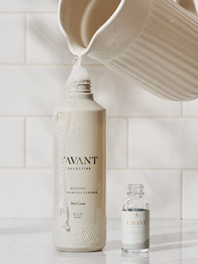 Porclein pitcher filling a 16oz Multipurpose tan PCR bottle with suds/cleaner flowin over. Empty 1oz concentrate bottle to the right. White tile background.