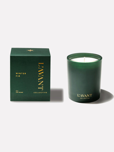 Green Winter Fir holiday scented candle with gold foil L'AVANT Collective logo at base. Green retail box to the left of the candle.
