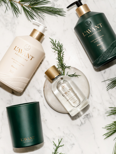 Four Products displayed laying down on marble counter, Winter Fir Green Candle, Winter Fir Dish Soap Cream Color Glass Bottle, Winter Fir Hand Soap Green Bottle, Winter Fir Room Spray in Clear Glass bottle with Gold cap,Greenery in background.