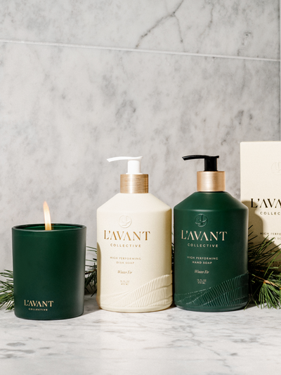 Winter Fir Green Candle lit with Flame, Winter Fir Dish Soap Cream Color Glass Bottle, Winter Fir Hand Soap Green Bottle, Marble Counter and Backdrop with Greenery in background.