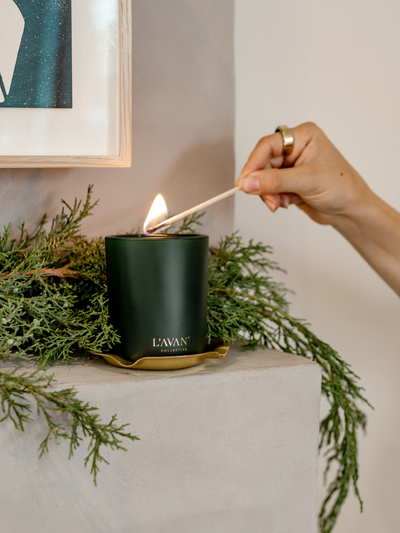 Green Winter Fir Candle with gold foil L'AVANT Collective logo at base. Hand is lighting holiday candle with match.