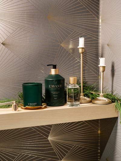Three holiday Products displayed, Winter Fir Green Christmas scent Candle with lit flame, Winter Fir Hand Soap Green Bottle, Winter Fir holiday scented Room Spray in Clear Glass bottle with Gold cap, with Greenery in background and Gold Candlesticks.