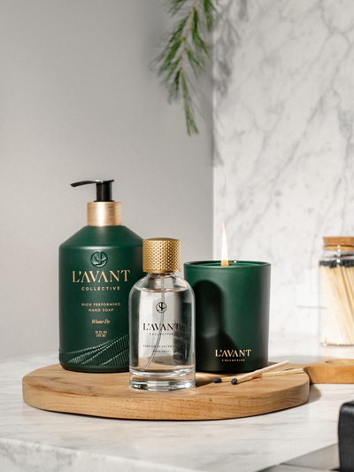 Three Products displayed, Winter Fir Green Candle with lit flame, Winter Fir Hand Soap Green Bottle, Winter Fir Room Spray in Clear Glass bottle with Gold cap,Greenery in background.