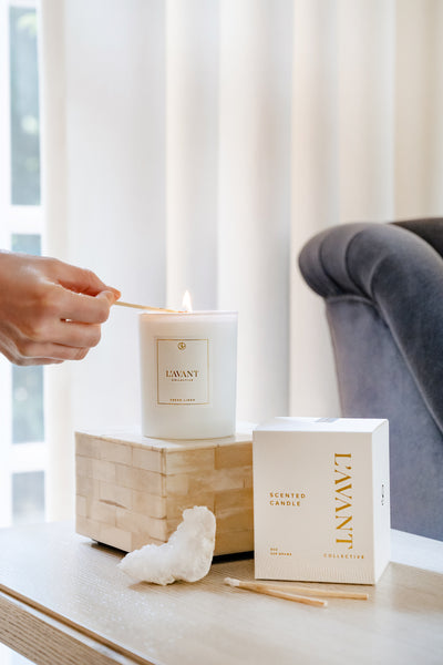 White Fresh Linen Candle in custom glass, 10 oz white candle jar with white candle inside. White L'AVANT sticker with gold writing on the candle jar.  Hand to the left, reaching in to lite candle sitting on design box. below small piece of white crystal and to the right, white L'AVANT candle box with gold foil text and two match sticks