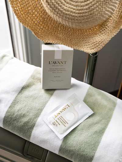 A stylish travel-inspired scene featuring L'AVANT Collective High-Performing Laundry Detergent Travel Pouches in Fresh Linen scent. The elegant cream and gold packaging is displayed on a soft green and white striped towel, resting atop an olive green BEIS suitcase. A woven straw hat hangs above, evoking a chic and effortless travel aesthetic. This image highlights the convenience and sophistication of L'AVANT’s travel-friendly detergent, perfect for on-the-go freshness with a touch of luxury.