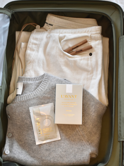 An open suitcase packed with neutral-toned travel essentials, including a folded gray cashmere sweater, white denim jeans with "Still Here" branding, and two Rhode lip products tucked into the pocket. L'AVANT Collective High-Performing Laundry Detergent Travel Pouches in Fresh Linen scent are neatly placed on top, showcasing their elegant cream and gold packaging. The scene exudes a sense of luxury, convenience, and effortless style—perfect for maintaining fresh, clean clothes while traveling.