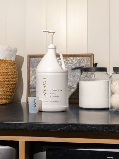 Image of a laundry room countertop featuring a one-gallon jug of L'AVANT Collective High Performance Laundry Detergent labeled 'Fresh Linen, 5x Concentrated' with a pump dispenser. Next to the jug is a small measuring cup with the L'AVANT logo. The countertop also includes a glass jar of powdered detergent and another jar with wool dryer balls. In the background, a woven basket filled with folded white towels and a decorative metallic framed artwork add aesthetic touches.