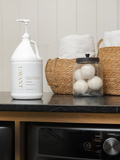 Image of a laundry room countertop featuring a one-gallon jug of L'AVANT Collective High Performance Laundry Detergent labeled 'Fresh Linen, 5x Concentrated' with a pump dispenser. The detergent sits on a dark marble counter next to a clear jar containing wool dryer balls and woven baskets filled with neatly folded white towels. Below the counter, the control panel of a washing machine is visible.