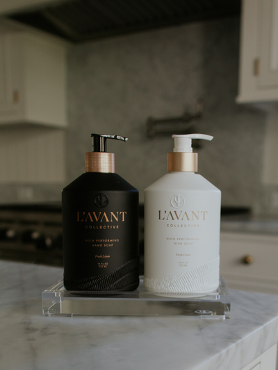 Custom glass, 16 oz black bottle with hand soap inside. White custom glass 16oz bottle wtih gold foil text "L'AVANT Collective" brushed gold neck on white pump with dish soap inside. . Clear lucite tray with LC logo etched in bottom right corner. The two bottles are sitting on the tray which is on a marble kitchen countertop.