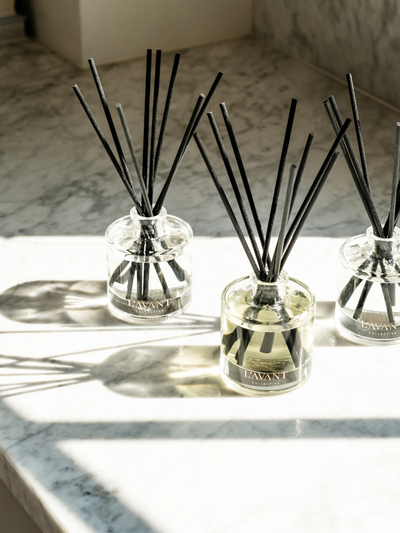 Multiple reed diffusers with black reeds are grouped together on a marble surface. Natural light streams through the space, casting elegant shadows and highlighting the glass vessels' reflections.