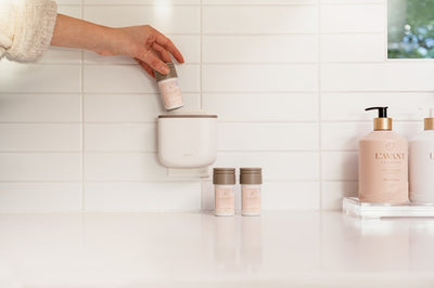 hand holding a pink area pod and placing into white aera diffuser with two pink pods on white countertop.  Pink glass bottle on lucite tray to the right.