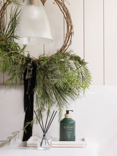 Christmas scented hand soap on a marble tray next to holiday fragranced oil and reeds in a glass jar. Christmas decorations in the background.