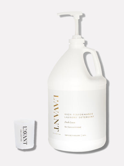 Image of a L'AVANT Collective product. A white one-gallon jug of high-performance laundry detergent labeled 'Fresh Linen, 5x Concentrated.' The jug features a sleek, minimalistic design with gold lettering and a pump dispenser. Next to the jug is a small measuring cup with the L'AVANT Collective logo printed in black.