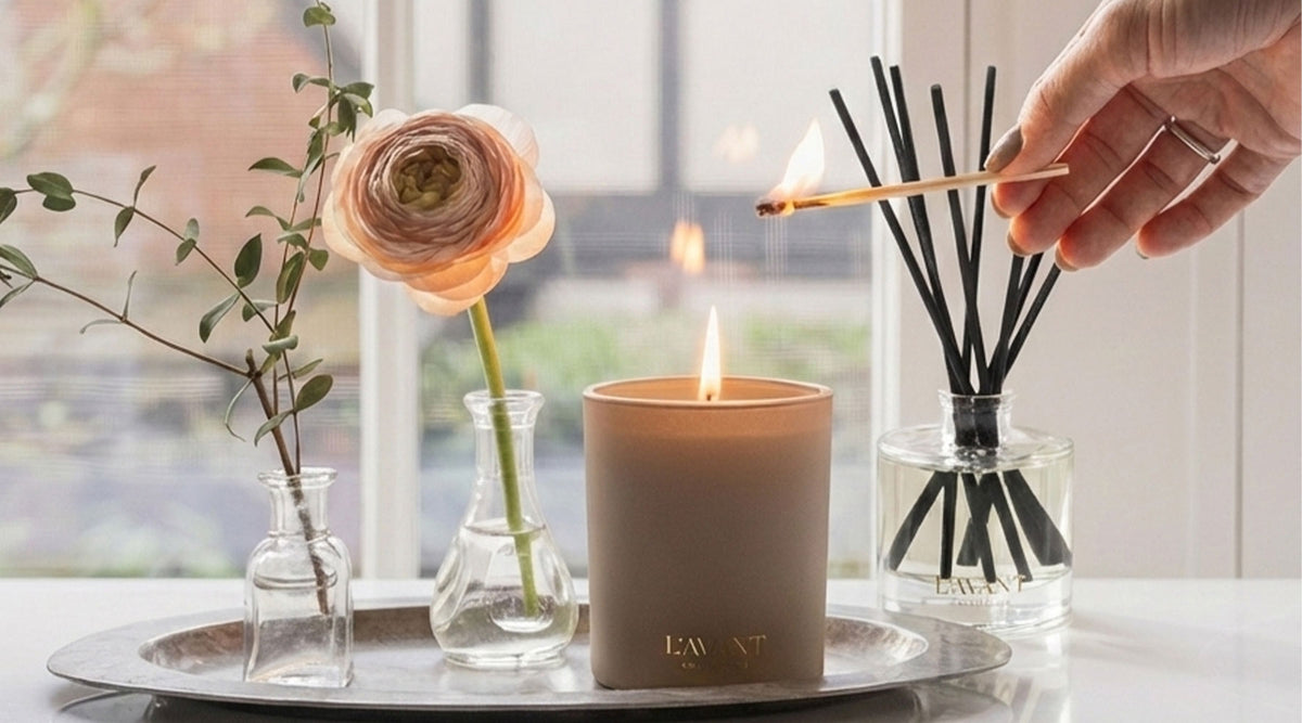 A candle is being lit in front of a reed diffuser on a countertop with some flowers.