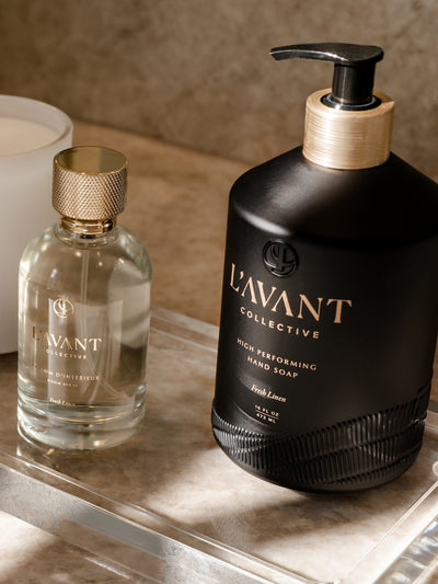 Clear glass bottle with L'AVANT logo stamped in gold foil. Glass bottle with gold topper. Sitting next to the room spray is a black custom, glass bottle of hand soap with a gold neck and black pump for the soap.