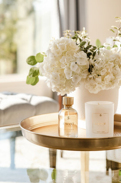 Clear glass bottle of room spray with gold topper sitting on side table. Next to the room spray bottle is a white glass candle and a bouquet of fresh white flowers in the background.