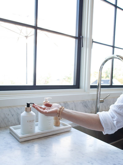 Custom white bottle of lotion with white bottle of dish soap on marble tray. Model is holding hand out to lotion. All products on a marble tray by a sink.