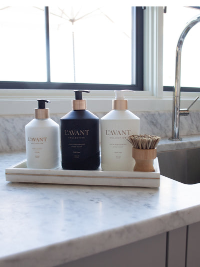 Sink side starter bundle. 10oz glass bottle of hand lotion and white bottle of dish soap and black bottle of hand soap and wood dish scrubber with marble tray.  Image is a lifestyle image with a marble background in front of a window and by a sink.