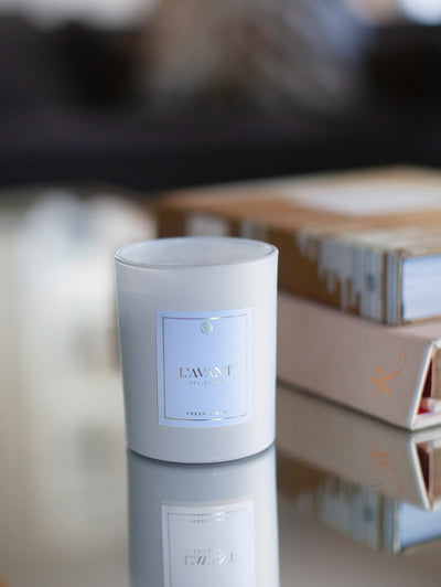 Linen Scented Candle White Fresh Linen Candle in custom glass, 10 oz white candle jar with white candle inside. White L'AVANT sticker with gold writing on the candle jar. On mirrored surface with two books in blurred background